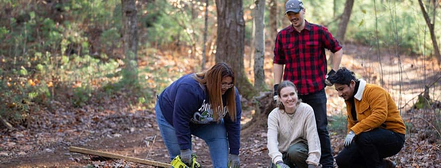 Two female and a male student crouching as well as one male student standing in a wooded area working on a sustainability project