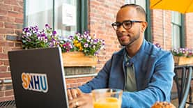 Geremy Laboy Prospere, sitting at an outdoor cafe working on a laptop with an SNHU sticker on the cover.