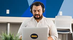 Jesus Suarez, who earned his bachelor's degree in graphic design and media arts in 2021, wearing headphones and typing on a laptop that has an SNHU sticker on the cover.